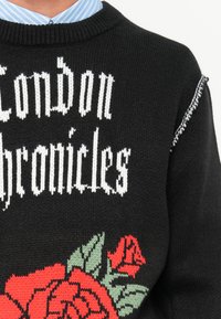 Zwarte gebreide trui met "London Chronicles" in witte tekst. Versierd met rode en groene rozen, afgewerkt met een geribbelde halslijn.