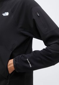 Čierny rukáv bundy North Face s vreckom na zips na hornej časti ramena a textom "FUTUREFLEECE" blízko zápästia, ruka vo vrecku na boku.