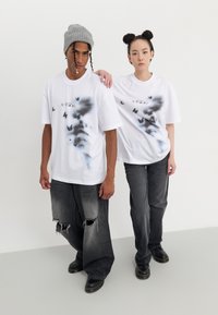 Camisetas blancas de gran tamaño con un gráfico de mariposa negra y el texto "VISION", combinadas con jeans grises desgastados y calzado negro.
