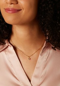 Gouden ketting met een ronde hanger met een heldere edelsteen. De ketting ligt op een zijden blouse met een subtiele glans.