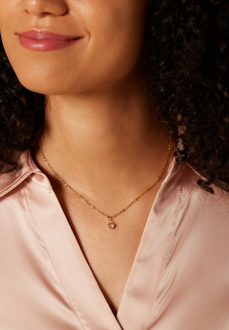Gouden ketting met een ronde hanger met een heldere edelsteen. De ketting ligt op een zijden blouse met een subtiele glans.