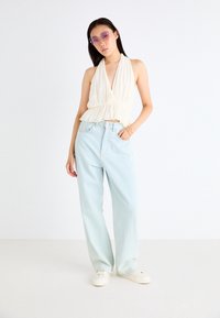 Hvit halterneck-top med samlet tekstur, kombinert med lyseblå høytlivsjeans og hvite sneakers. Modellen har på seg lilla solbriller.