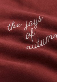 Bordová tkanina s bielym výšivkovým nápisom „radosťou jesene“ a s jemným textúrovaným povrchom.