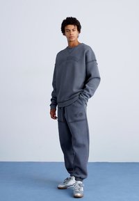 Grå hettegenser og matchende joggebukser i blanding av bomull. Hettegenseren har ribbestrikkede mansjetter og logo detalj, mens buksene har sidelommer. Sølvfargede sneakers.