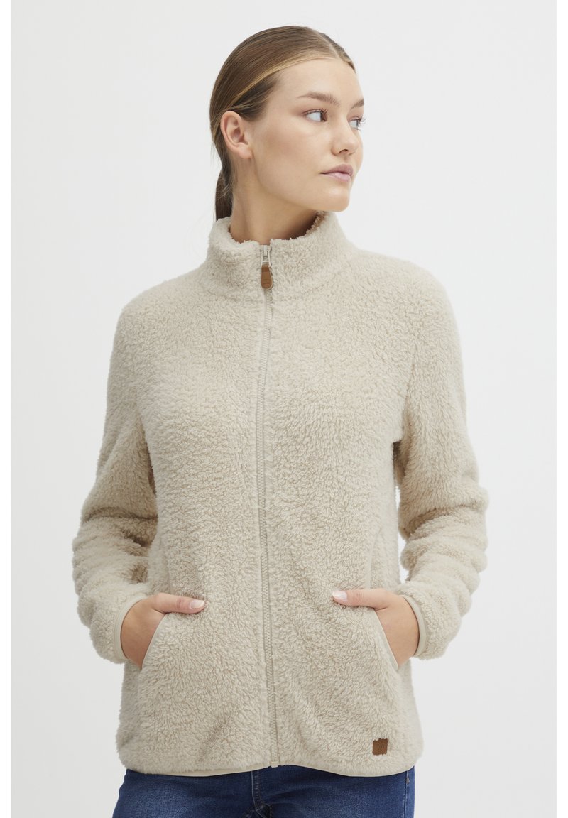 Beige fleecejakke med høy krage, hel glidelås og sidelommer. Har teksturert stoff og en liten logodetalj nederst.