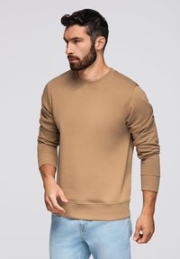Felpa color sabbia con collo a giro, polsini e orlo a costine. Il tessuto appare morbido e confortevole. Modello che indossa jeans azzurri.