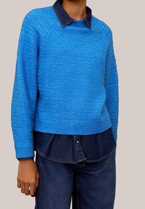Personne portant un pull bleu vif texturé superposé à une chemise en denim bleu foncé et un jean en denim bleu foncé assorti.