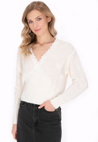 Maglione bianco a maglia con scollatura a V profonda e bordo ondulato. Indossato sopra una gonna in denim nera, evidenziando un look casual a strati.