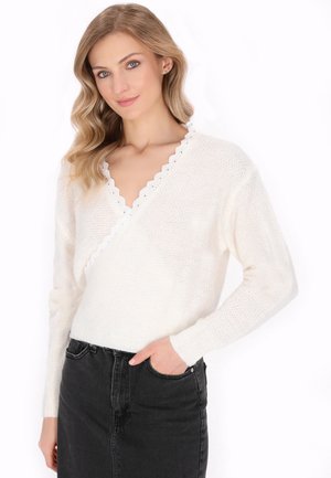 Pull en tricot blanc avec un décolleté en V profond, orné d'un ourlet en dentelle. Porté sur une jupe en denim noire, mettant en valeur un look décontracté en couches.