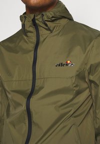 Giacca leggera verde oliva con zip frontale, cappuccio regolabile, dettagli della zip neri e un piccolo logo sul petto.