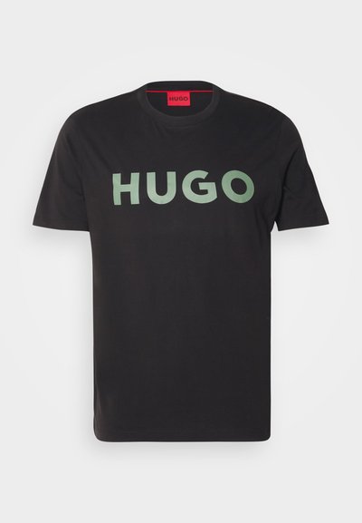 Черен памучен T-shirt с голям, смел, зелен логотип "HUGO" в централната част. Класическо кръгло деколте и къси ръкави. Обикновен фон.