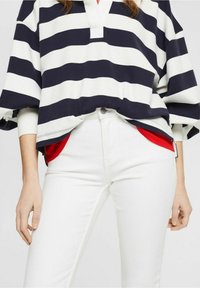 Námořnický a bílý pruhovaný oversized pulovr s výstřihem do V a širokými rukávy, spárovaný s úzkými bílými džíny a červeným podšívkovým kapsou.