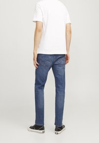 Vaqueros de mezclilla azul de corte ajustado, con dos bolsillos traseros que presentan detalles de costura, combinados con una camiseta blanca de manga corta y zapatillas negras.