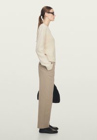 Beige gebreide trui, hoogliggende beige broek, zwarte schoenen en een donkere tas. Zijaanzicht tegen een neutrale achtergrond.