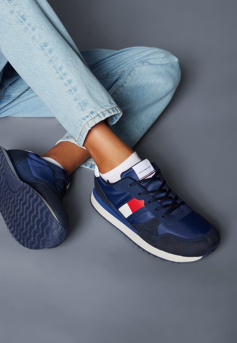 Personne portant un jean bleu clair et des baskets bleu marine avec un logo rouge et blanc assise sur un sol gris, jambes croisées.