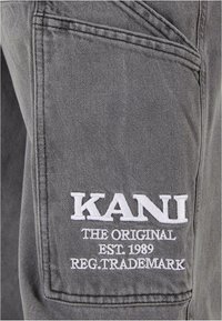Tela gris con un tejido texturizado que presenta un bolsillo cosido con bordado blanco que dice "KANI THE ORIGINAL EST. 1989 REG. TRADEMARK."