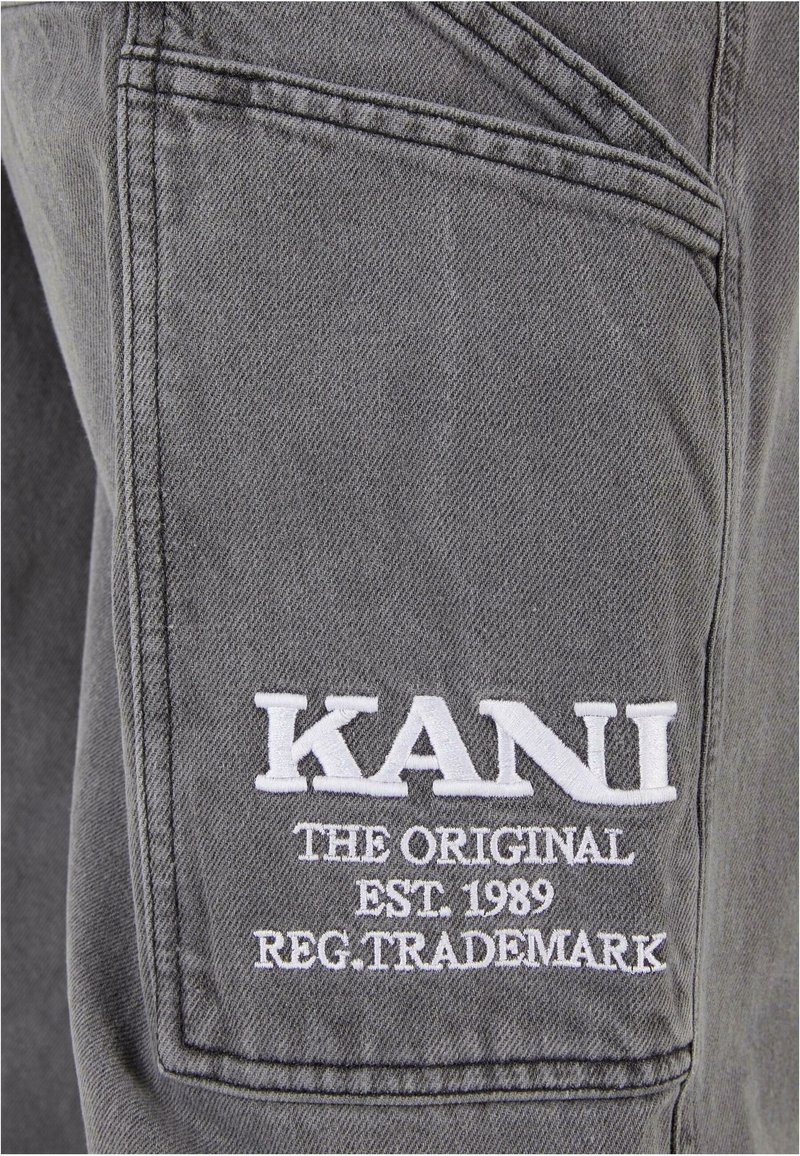 Un tissu gris avec un tissage texturé présente une poche cousue affichant une broderie blanche portant les mots "KANI L'ORIGINAL EST. 1989 MARQUE DÉPOSÉE."