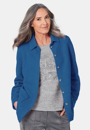 Cardigan en laine bleu, boutonnière, avec un col et deux poches. Porté par-dessus un pull en gris clair avec un motif texturé.