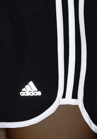 Bermudas negras de Adidas con ribetes blancos y tres franjas blancas a lo largo del lado, mostrando parte del muslo de una persona.