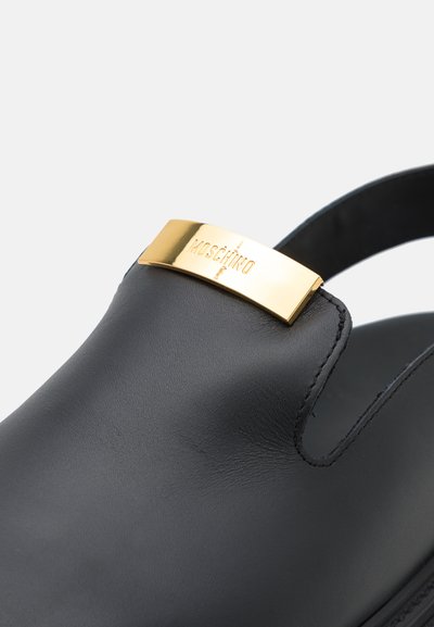 Melnas ādas kurpes ar noapaļotu purngalu un zelta metāla siksniņu, kas rotāta ar "Moschino" logotipu. Gluda tekstūra, minimālisma dizains un atvērta papēža daļa.