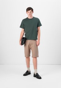T-shirt en coton vert, shorts beige à côtes, mocassins noirs et chaussettes claires ; mannequin tenant une pochette noire. Style simple et décontracté.