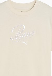 Bežs kokvilnas T-krekls ar maigu tekstūru, kurā ir sudraba izšūts "Puma" logo un mazs Puma logo uz kakla izgriezuma.