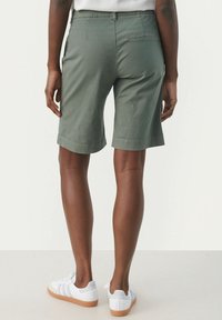 Shorts verts en coton à coupe droite, avec des ourlets roulés et une poche arrière. Portés avec des baskets blanches, montrant une tenue décontractée.