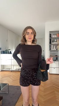 Bluză neagră cu mâneci lungi și manșete evazate, asortată cu pantaloni scurți închiși la culoare cu paiete. Ținuta este completată de o geantă neagră strălucitoare.