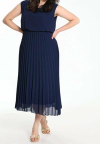 Robe plissée bleu marine avec un design sans manches et un corsage ajusté, dotée d'une jupe fluide atteignant le milieu du mollet, complétée par des sandales beiges.