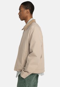 Chaqueta beige con cuello de pana, bolsillos laterales y un corte relajado. La tela parece suave, con un diseño minimalista y acentos sutiles.