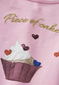 Stickerei auf pinkem Stoff mit goldener Schrift "Stück Kuchen", einer Illustration eines Cupcakes, der mit weißer Glasur und bunten Herzformen verziert ist.