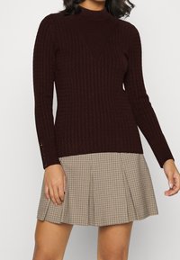 Maglione ribbed di un intenso bordeaux con colletto alto, caratterizzato da un motivo testurizzato e dettagli con bottoni sulle maniche, abbinato a una gonna a quadri.