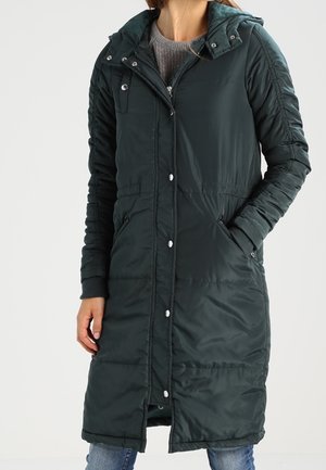 Femme portant un long manteau d’hiver matelassé vert foncé avec des boutons devant et des poches latérales zippées, par-dessus un pull gris et un jean.
