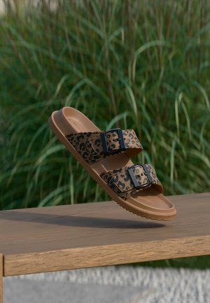 Sandale marron avec des brides à imprimé léopard noir et des boucles noires flottant au-dessus d'un banc en bois en plein air, avec un fond d'herbe verte.