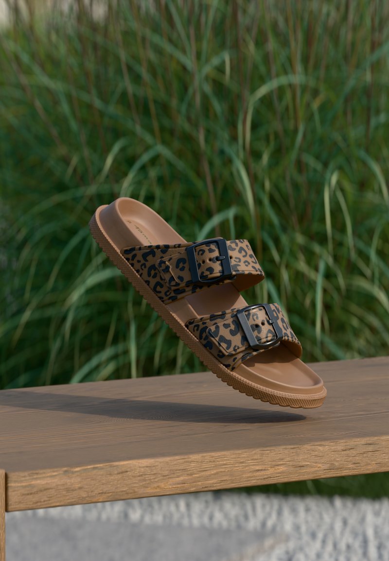 Καφέ σανδάλι με μαύρα λουριά με λεοπάρ σχέδιο και μαύρα αγκράφες, επιπλέει πάνω από ξύλινο παγκάκι σε εξωτερικό χώρο με φόντο πράσινο γρασίδι.