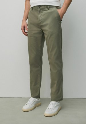 Mann trägt olivgrüne gerade geschnittene Hosen und weiße Turnschuhe, steht mit einer Hand in der Tasche vor einem einfarbigen Hintergrund.