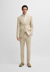 Beige tailored suit with notch lapels, single-breasted jacket, and slim-fit trousers. Paired with a white shirt and light brown shoes.