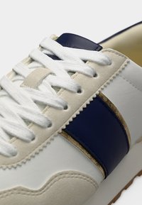 Κοντινό πλάνο σε λευκό αθλητικό παπούτσι με μπεζ σουέντ λεπτομέρειες, ναυτικό μπλε δερμάτινη λωρίδα και λευκά κορδόνια που περνούν μέσα από εσωτερικά τρύπες.