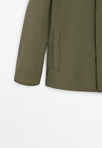 Giacca leggera verde oliva con taglio dritto, maniche lunghe, tasche laterali e una texture liscia con dettagli di cucitura minimi.