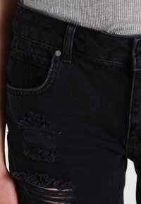 Zwarte gescheurde spijkerbroek met rafelige stukken en een klassiek ontwerp met vijf zakken. Heeft een metalen knoopsluiting en een gestructureerde stof.