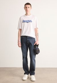 White short-sleeve t-shirt with blue printed "Hugo" logo, dark blue jeans, white sneakers, holding a small black bag with blue accent.