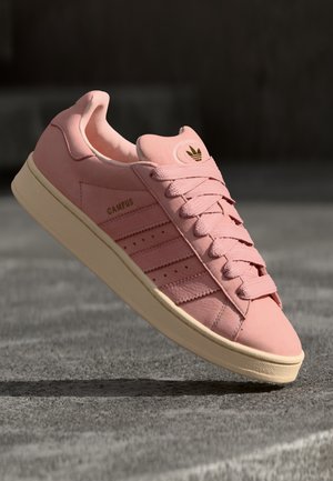 Růžová teniska Adidas Campus s béžovou podrážkou a tkaničkami, zobrazující logo a pruhy, umístěná na šedém betonovém povrchu.