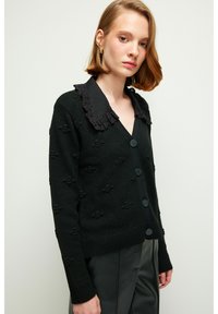 Svart stickad cardigan med texturerat blommönster, V-ringning och krage med spetsdetaljer. Har stora svarta knappar och ribbade ärmslut.