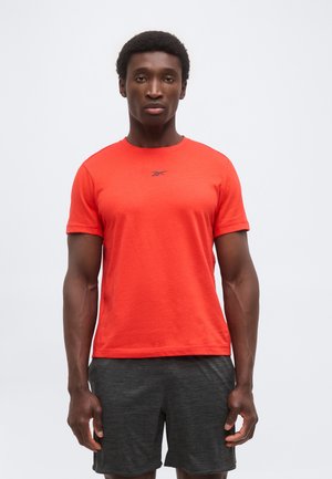 Uomo con maglietta sportiva arancione brillante a maniche corte e pantaloncini grigio scuro, in piedi di fronte a uno sfondo semplice.