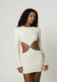Frau mit lockigem schwarzen Haar, die ein cremefarbenes, langärmliges Spitzenkleid mit seitlichen Ausschnitten und einem dekorativen muschelförmigen Verschluss in der Taille trägt.