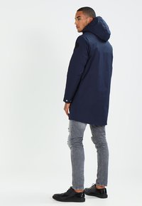 Cappotto impermeabile blu navy con cappuccio, caratterizzato da una vestibilità su misura, bottoni bianchi e toppe nere sulle spalle, abbinato a jeans grigi chiari e scarpe nere.