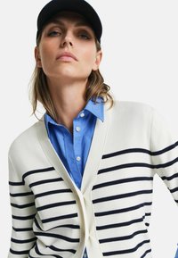 Cardigan rayé beige avec des lignes navy sur une chemise à boutons bleue. Le cardigan présente un décolleté en V et des boutons sur le devant.