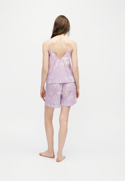 Femme debout pieds nus, de dos, portant un haut sans manches à fleurs couleur violet clair et un short assorti sur fond blanc.
