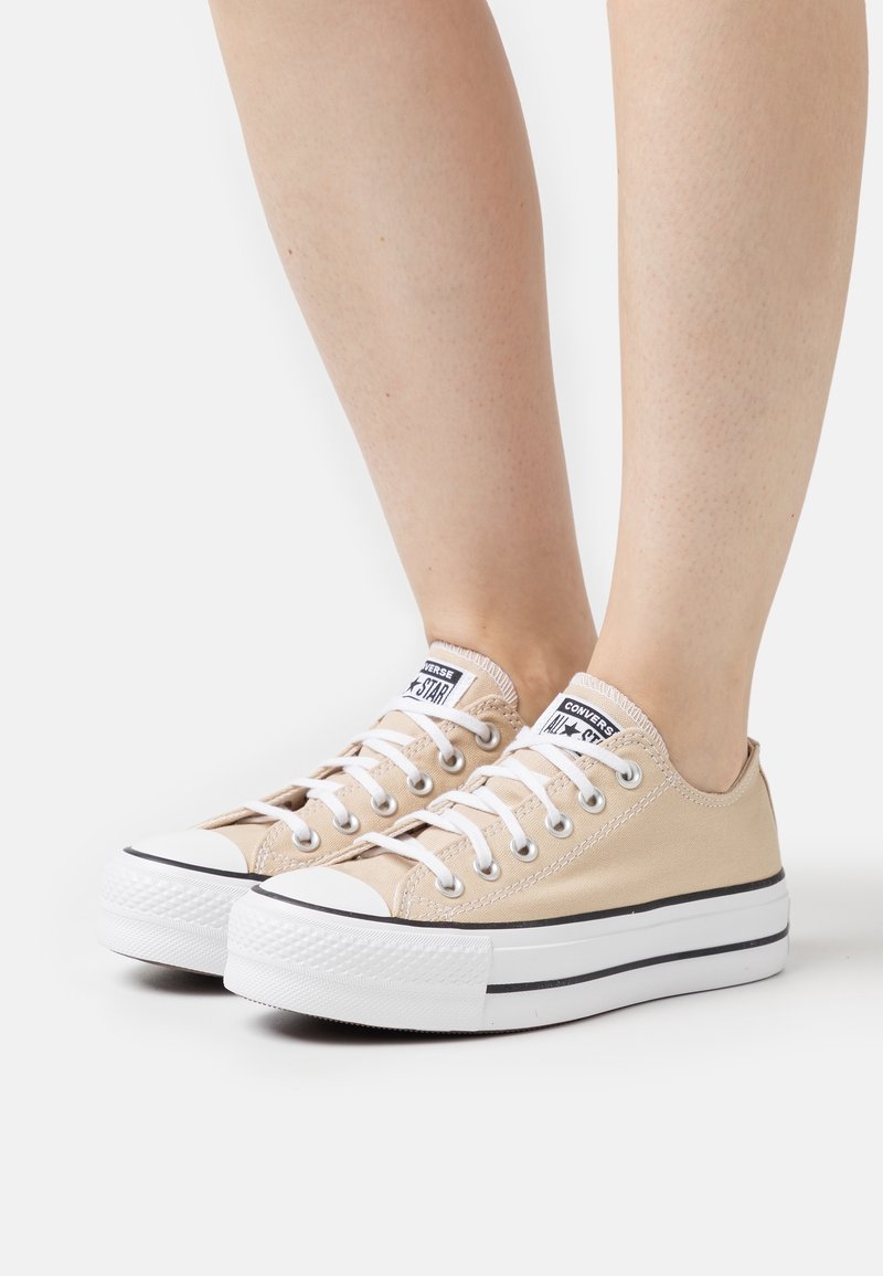 Baskets basses Converse All Star beiges avec des lacets blancs et des semelles épaisses blanches en caoutchouc portées sur des jambes nues.