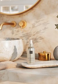 Zilveren spuitfles gelabeld "Leave-In Anti Frizz Conditioner" op een ronde marmeren schaal, met een beige handdoek en verschillende textuurrijke decoratieve items in de buurt.